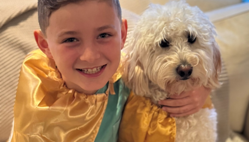 A young boy wears a gold cape while hugging his dog.