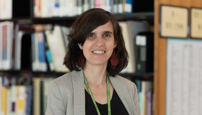 A young adult with light skin tone and short brown hair smiles and stands in front of library bookshelves.