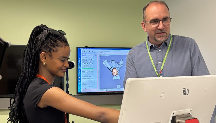 A female student is pointing to a screen next to a male adult who is standing