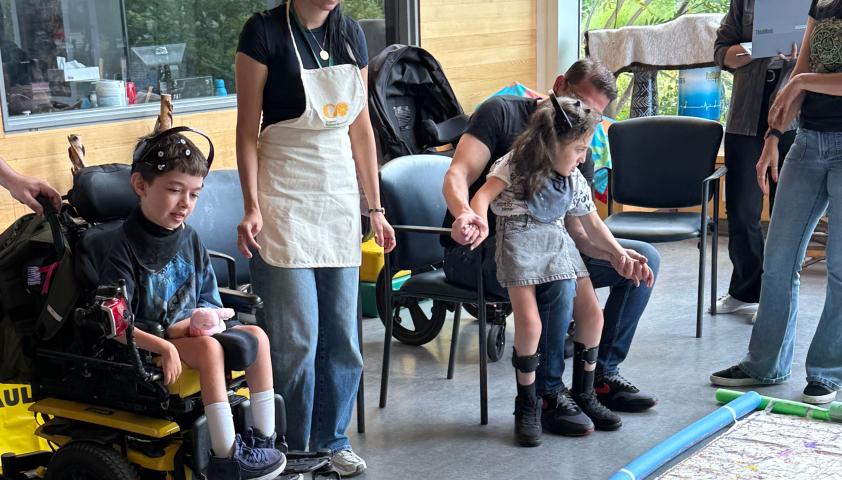 Two children are painting together. The children are wearing BCI headsets to control robotic balls dipped in acrylic paint. The painting is large and covers the floor. A clinician is standing beside the children. 