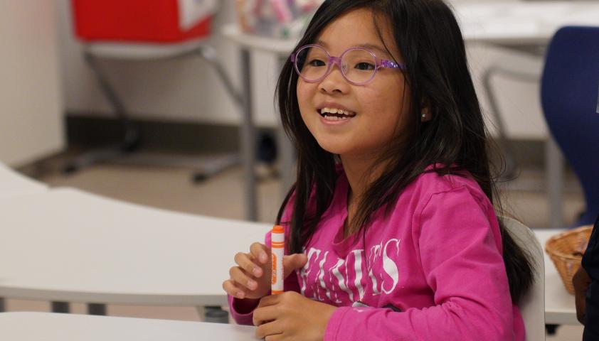 A young child smiles and holds a crayon