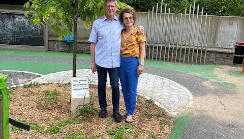 Two adults embrace outdoors beside a tall tree