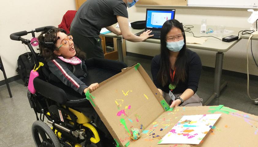 A child in a wheelchair wearing a BCI headset. The child is using the BCI to paint a picture with a ball. An adult is kneeling beside the child and holding a box with the painting inside of it.