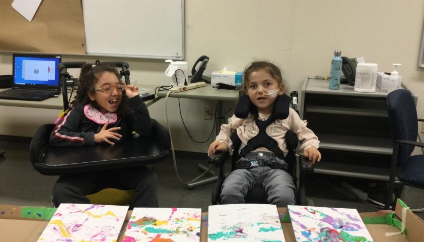 Two children in wheelchairs, with four colourful paintings on a table in front of them.