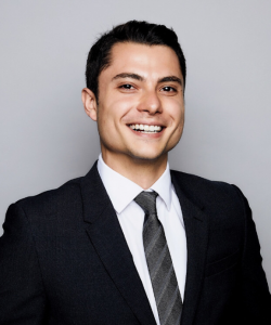 Dark-haired man smiling, wearing a black blazer and a grey, striped tie.