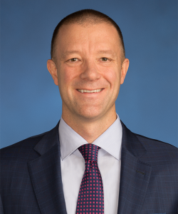 Man with brown, buzzed hair wearing a dark blue blazer, white shirt, and blue and red tie.