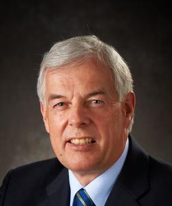 Man with gray hair wearing a dark blazer, light blue shirt, and a blue-striped tie