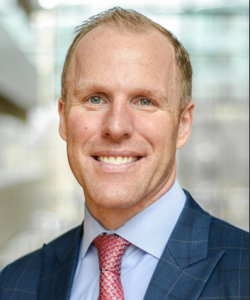 A smiling man with short blond hair and blue eyes. Wearing a dark blue plaid blazer, a light blue dress shirt, and a red patterned tie