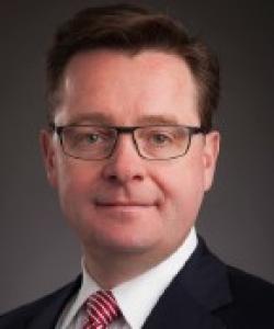 Man with short dark hair and black rectangular glasses. He's wearing a black blazer, white dress shirt, and a red and white striped tie