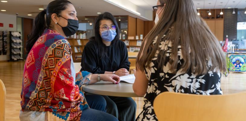 Three adults sit at a table together and have a conversation in the library.