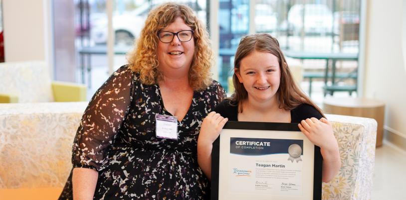 Dr. Carolyn Hunt and a child sitting beside each other on a couch. The child is holding up a framed certificate, and they are both smiling