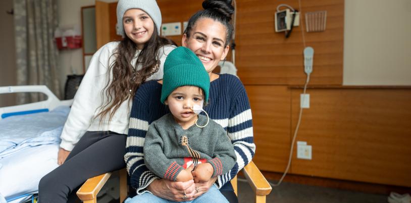 Two young children sit on their mom's lap, sitting on a chair in front of a hospital bed