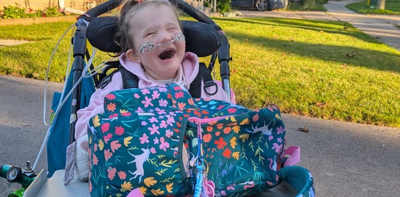 Young girl with oxygen prongs and huge smile sits in wheelchair with backpack on tray.