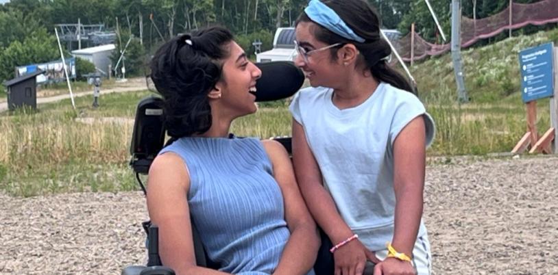 Teen girl in wheelchair and lavender outfit smiles at girl with bandana standing with arms on her chair