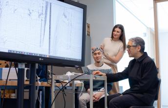 An adult sits and wears a brain computer interface headset while two other adults look at brain wave imaging.