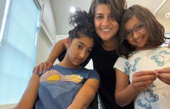 Woman in black shirt smiles with arms around teenage girl in wheelchair and young girl standing