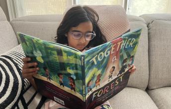 Girl with long dark hair and glasses reads large children's picture book