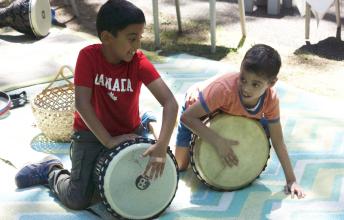 Boy with dark hair and red Canada shirt and boy with dark hair and orange shirt play drums