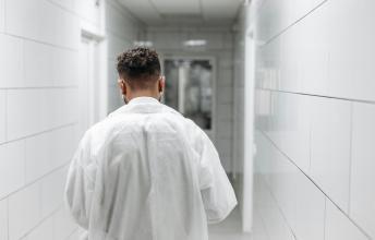 Back of man wearing white coat walking down hospital hallway.