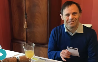 A man wearing a blue sweater sits at a table with a juice glass