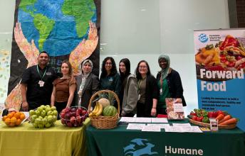 Photograph of seven people standing behind two tables displaying fresh fruits and vegetables at a Humane World for Animals event promoting sustainable, healthy, plant-based food systems. Tables feature green and yellow tablecloths, baskets of apples, bananas, and carrots, with a large poster and a backdrop of a painted Earth held by hands.