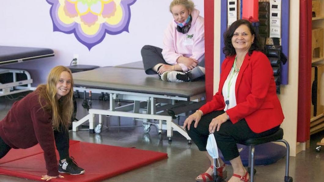 Woman on stool sitting in gym with two teen girls stretching on mats