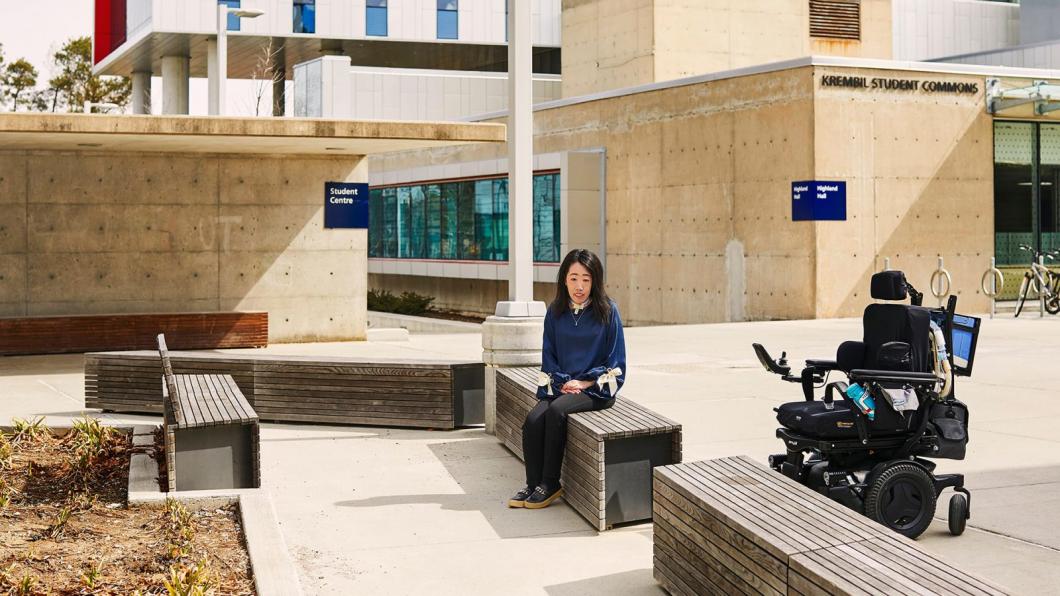 Young woman sits on bench in front of university with power wheelchair beside her