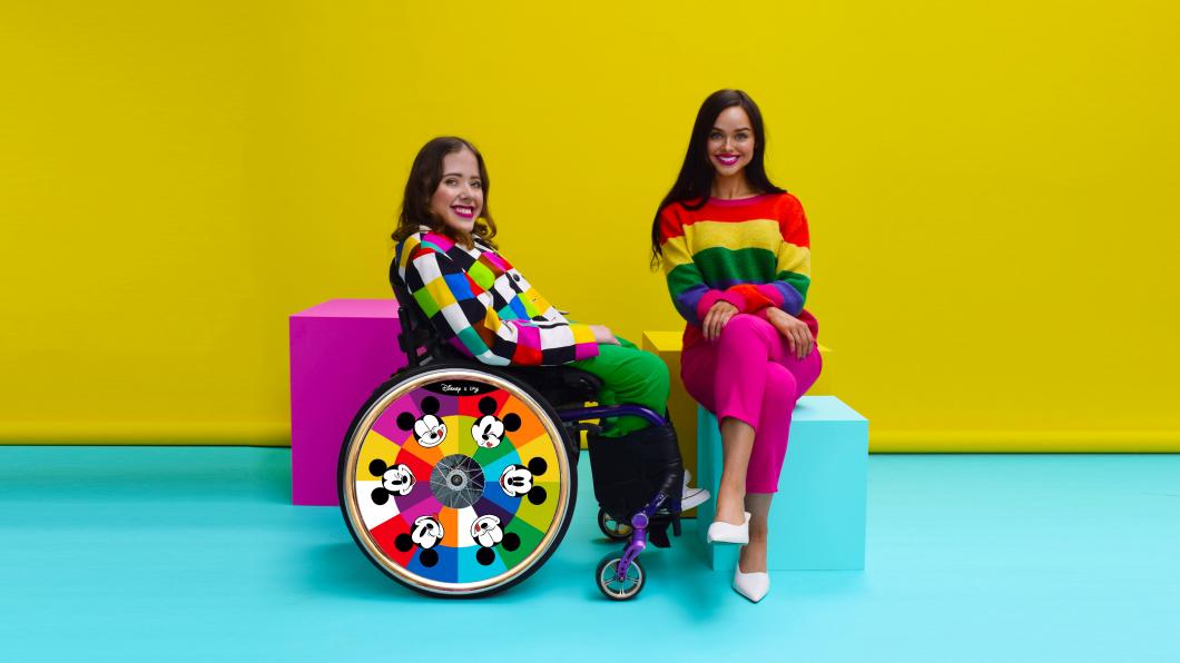 Two women in bright clothes, one in a wheelchair with a colourful wheelchair guard with Mickey Mouse on it