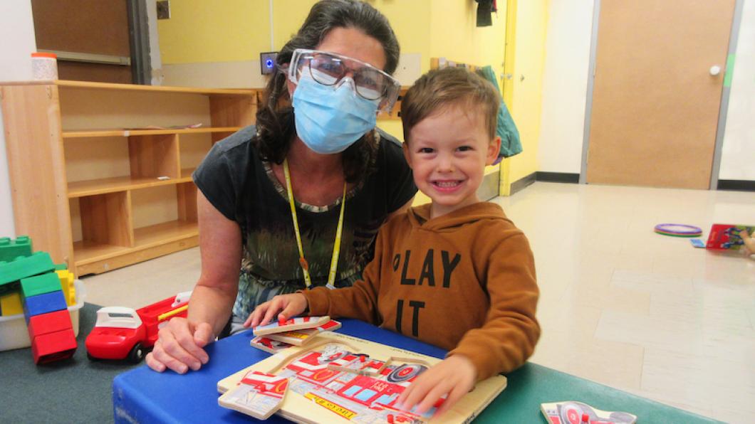 Woman with mask and face shield and goggles beside preschool boy doing a puzzle