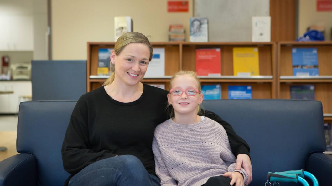 A mother and daughter sit together on a blue couch. The mother has blonde hair and wears a black sweater and blue jeans. The daughter has blonde hair and wears a grey sweater and pink glasses. Her mobility device can be seen at the corner of the image.