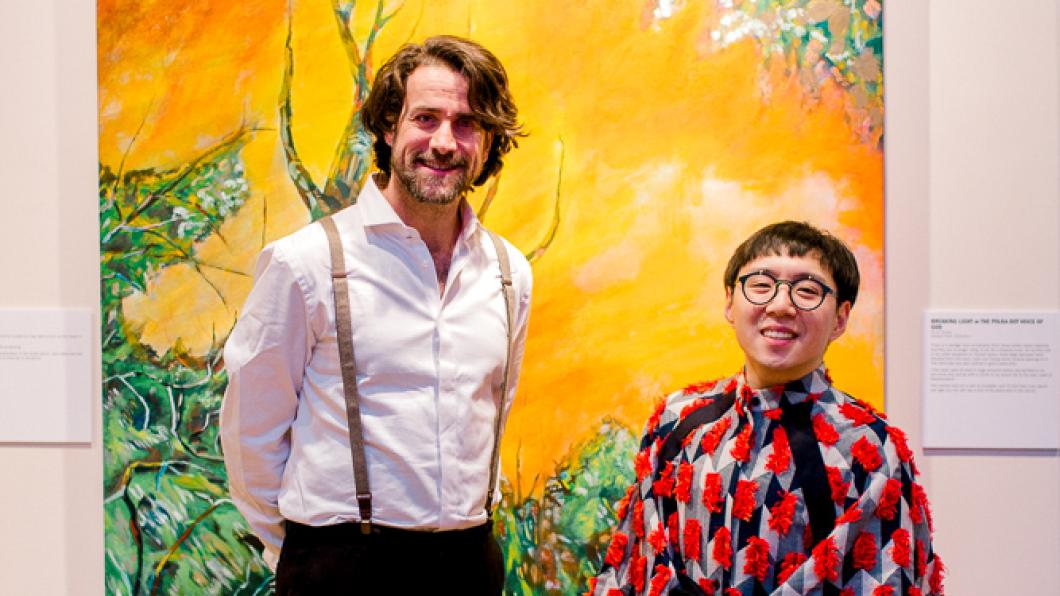 Bruce and Sean smile together at the opening reception for Through A Tired Eye. Sean is wearing a red kimono-like dresshirt with red fringes. Bruce wears a white dress shirt, black pants with grey suspenders. Behind them is an acrylic painting of greenery with a yellow skyline.