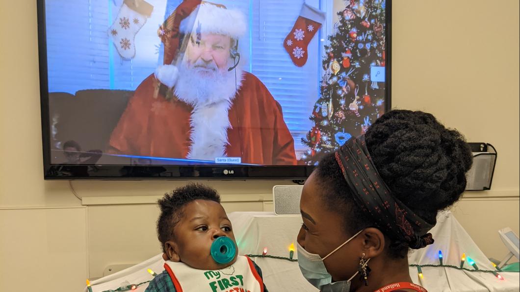 Parent, Julia, holding her child (and Holland Bloorview client), Thomas in front of a TV screen featuring Santa.