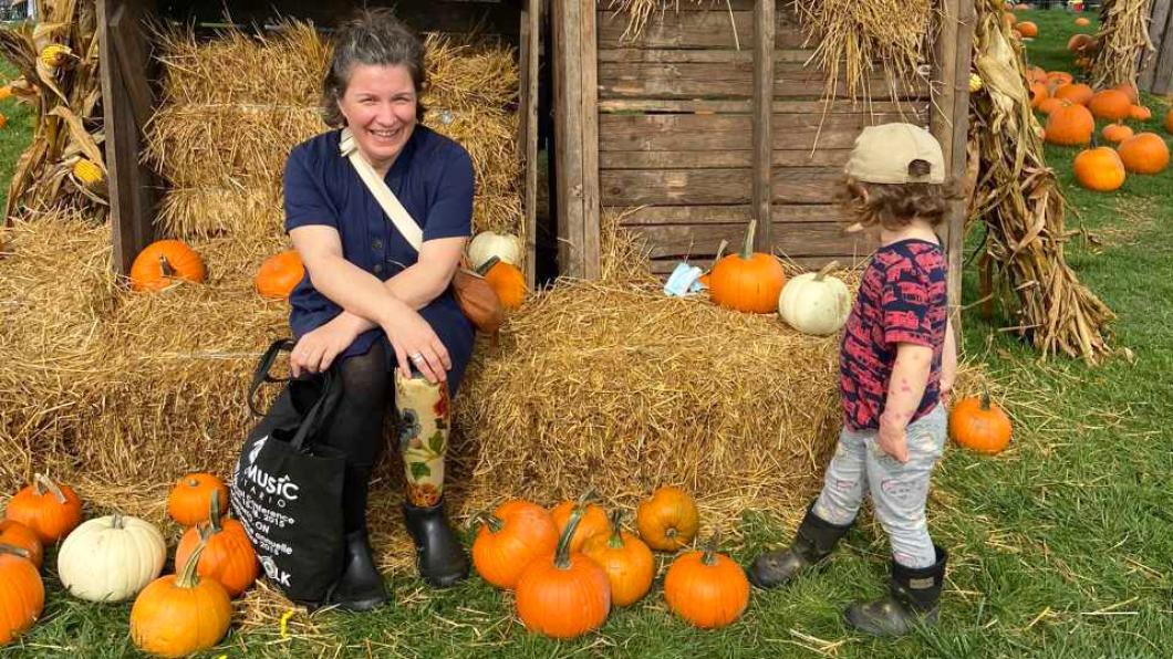 Woman on hay bail with flowers on prosthetic leg and small child nearby with pumpkins