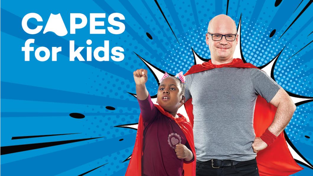 Image of adult man and younger male child posing as superheroes against blue backdrop that says "Capes for Kids"