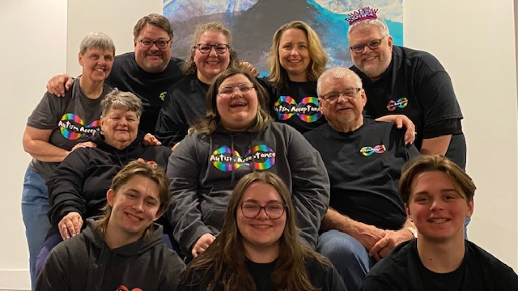 A smiling teen and their family sitting together for a family photo. Each family member is smiling and wearing black t-shirts and sweaters that say, "Autism Acceptance."