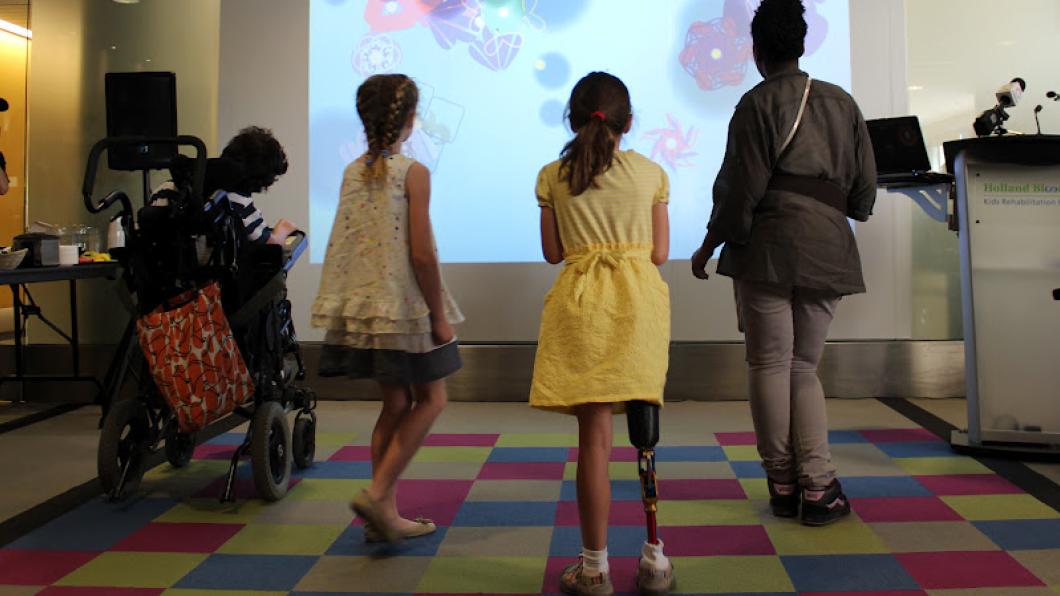 Children, two with disabilities, stand on coloured tiles in front of a screen with images on it.