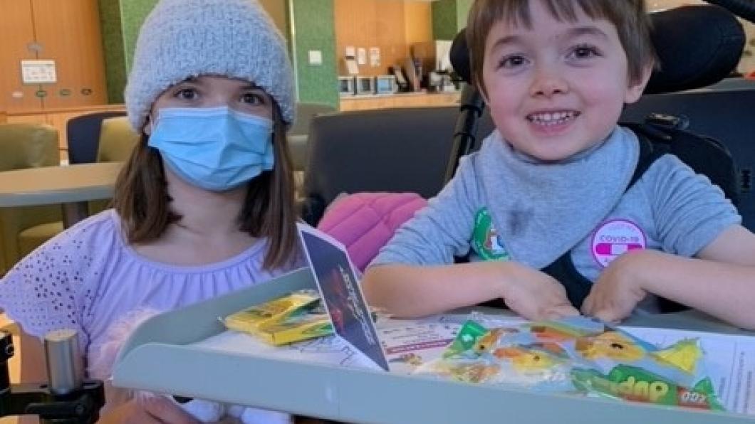 A young boy with light skin tone and short brown hair. He is using a wheelchair and a tray, which has school supplies and activities on it. A child with light skin tone, medium-length brown hair is kneeling beside him. They are wearing a knit hat and a blue face mask. 