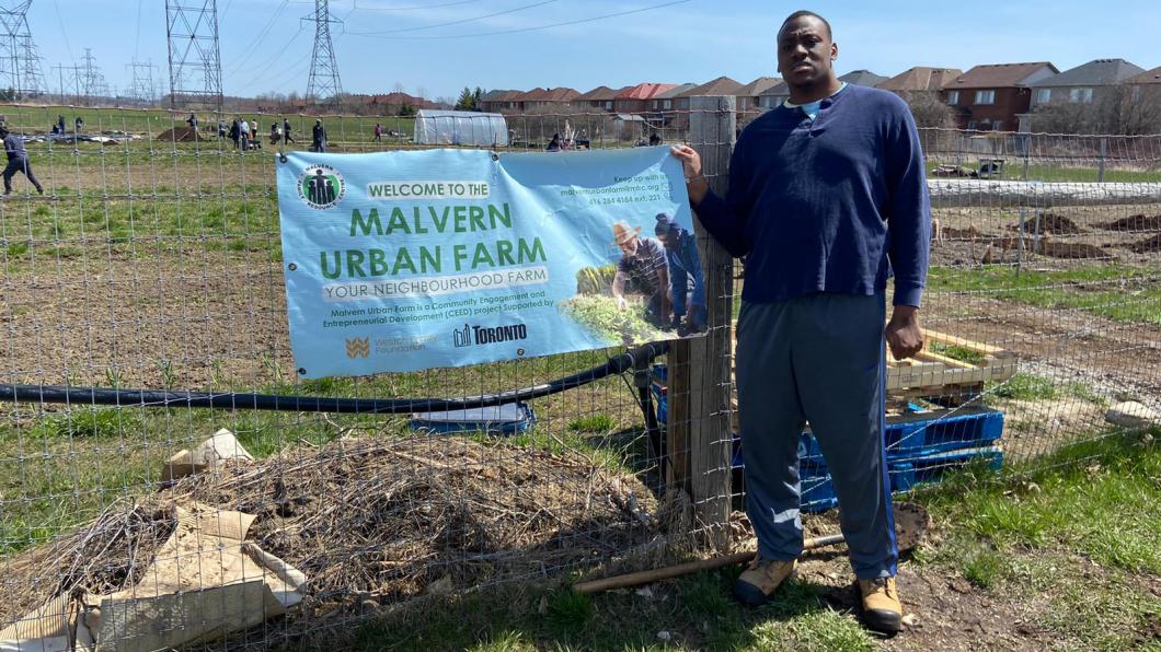 Young man stands in front of plots of land with banner for Malvern Urban Farm