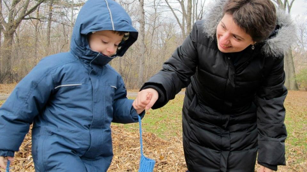 Woman holds hand of boy wearing blue winter coat with mittens on strings