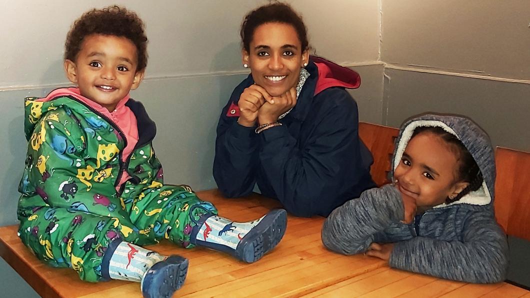 An adult sitting at a table with two children. They have deep skin tone, brown hair and are wearing a blue jacket. The child to their left is wearing a green snowsuit and rainboots with dinosaurs on it. The child on their right-hand side is wearing a grey sweater.