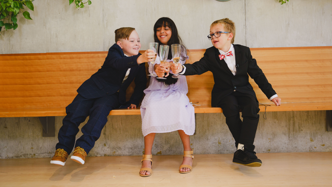 Three children sitting on a brown indoor bench. The first child has light skin tone, brown hair and is wearing a blue suit. The second child has medium skin tone, long black hair and is wearing a white dress. The third child has light skin tone, short blonde hair and is wearing glasses and a black suit. The children are toasting their drinks.