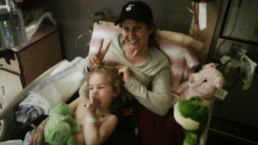 Toddler with blonde girls sucks thumb in hospital bed with mom wearing baseball camp