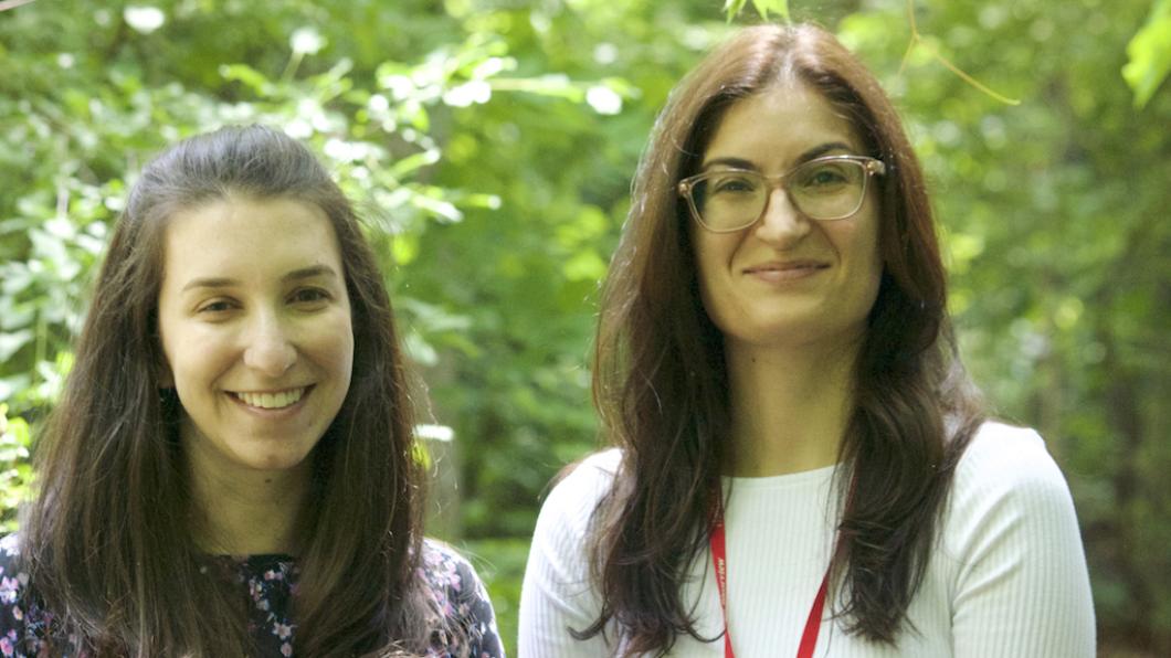 Two young women with long dark hair smile with trees behind them.