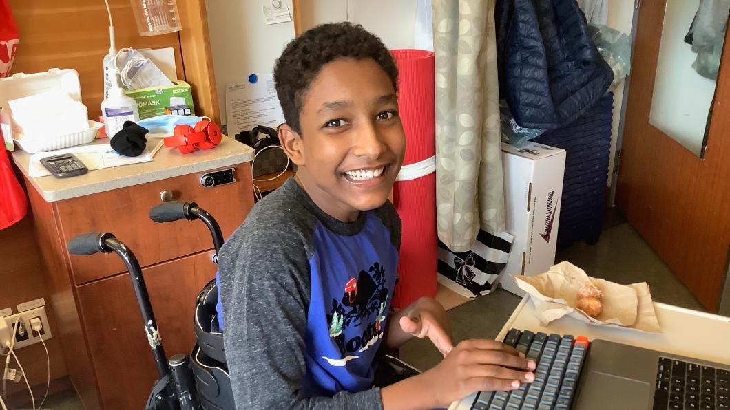 Teen with wide smile sits in wheelchair at computer in hospital room