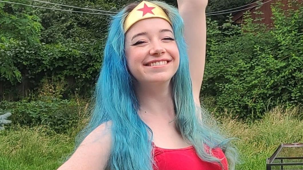 An adult with light skin tone and long blue hair wearing a gold crown-shaped hairband and a red tank top.