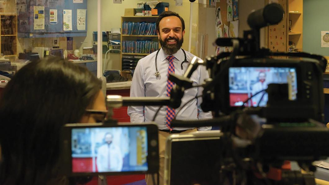 Bearded man with stethoscope around neck speaking in front of cameras