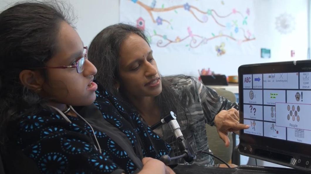 Teen wearing neckband and glasses sits with mother in front of computer screen