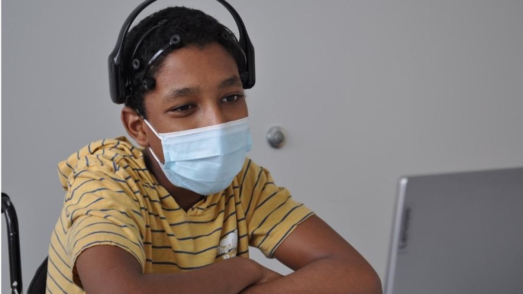 A boy with brown skin tone and black hair in a striped shirt, wearing a mask and headphones seated in front of a computer