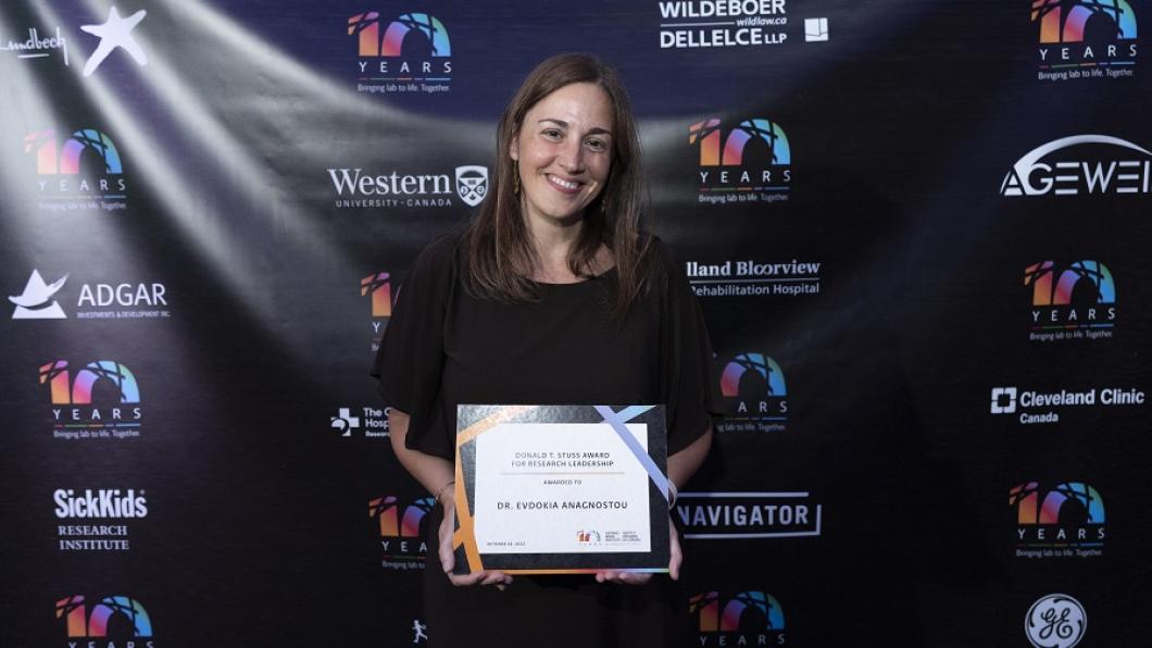 Dr. Evdokia Anagnostou stands holding her framed award, smiling. She is in front of a backdrop with various logos from Western, SickKids and other organizations.