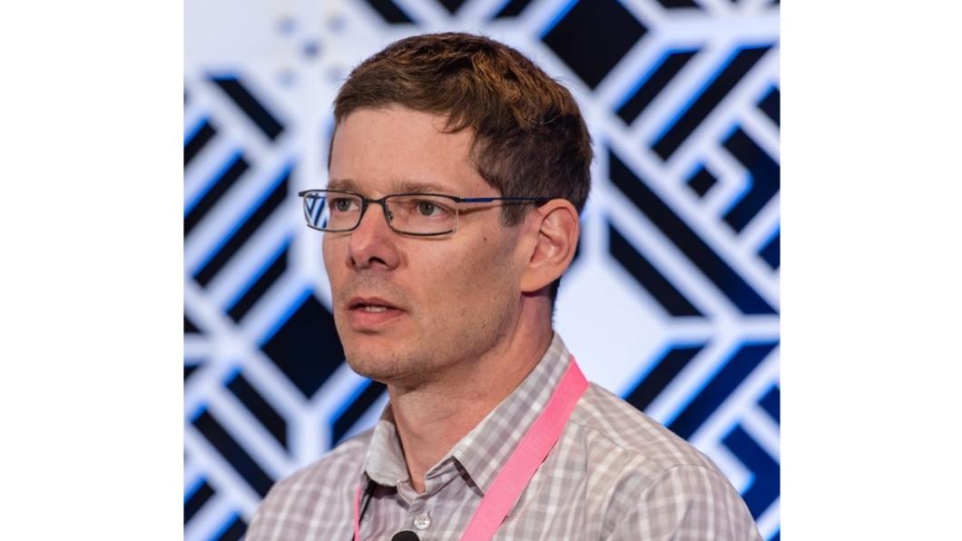 Dr. Jaimie Borisoff stands against a black and white checkered back drop wearing a light pink button up shirt, black glasses and short brown hair